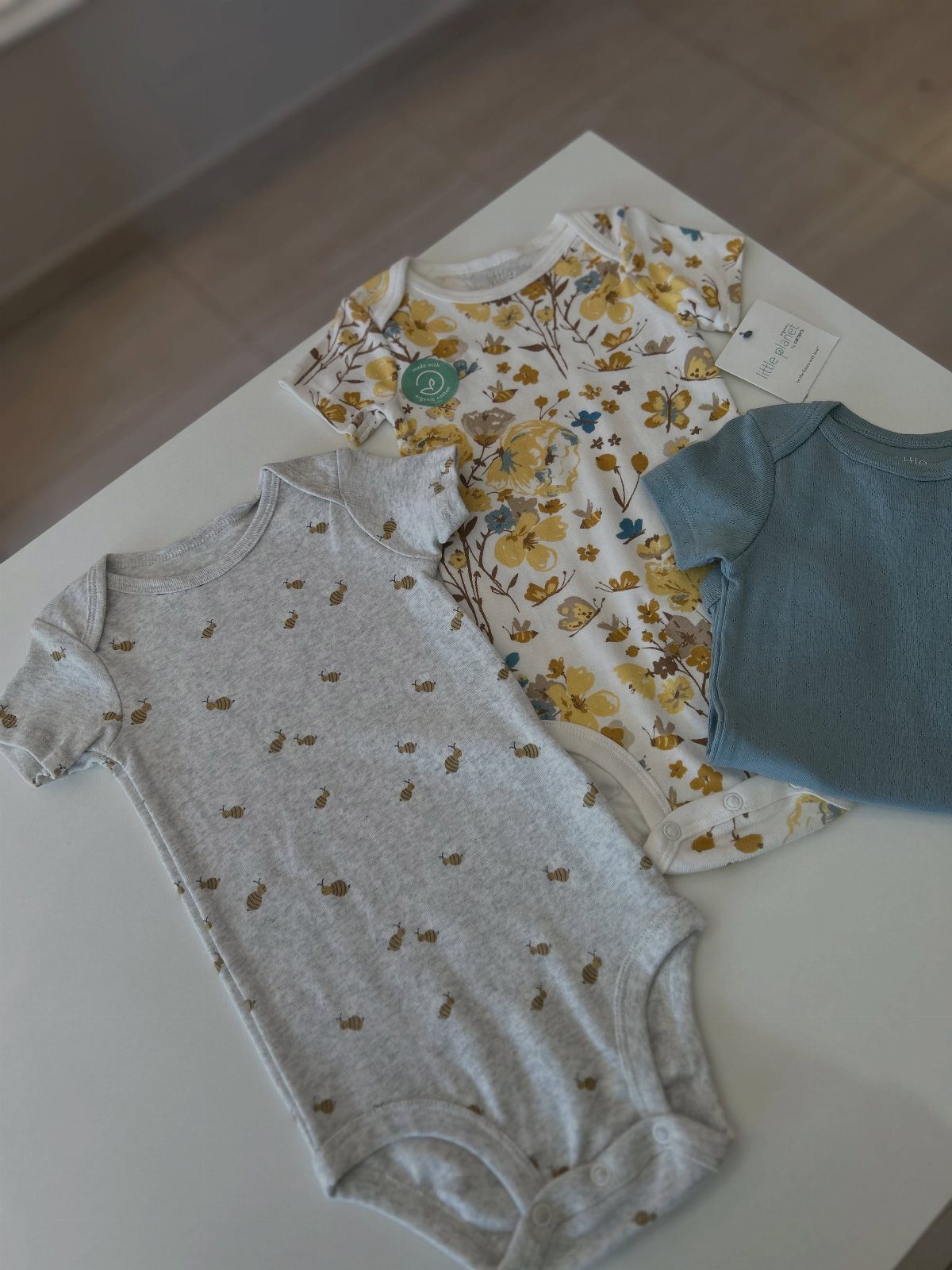 Three infant bodysuits in grey, floral, and blue laid on a white table.