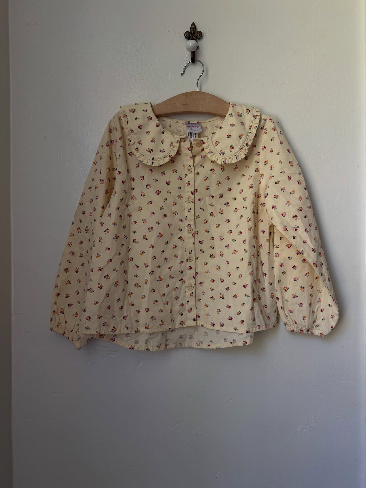 Cream floral infant blouse with Peter Pan collar hanging on a wooden hanger.