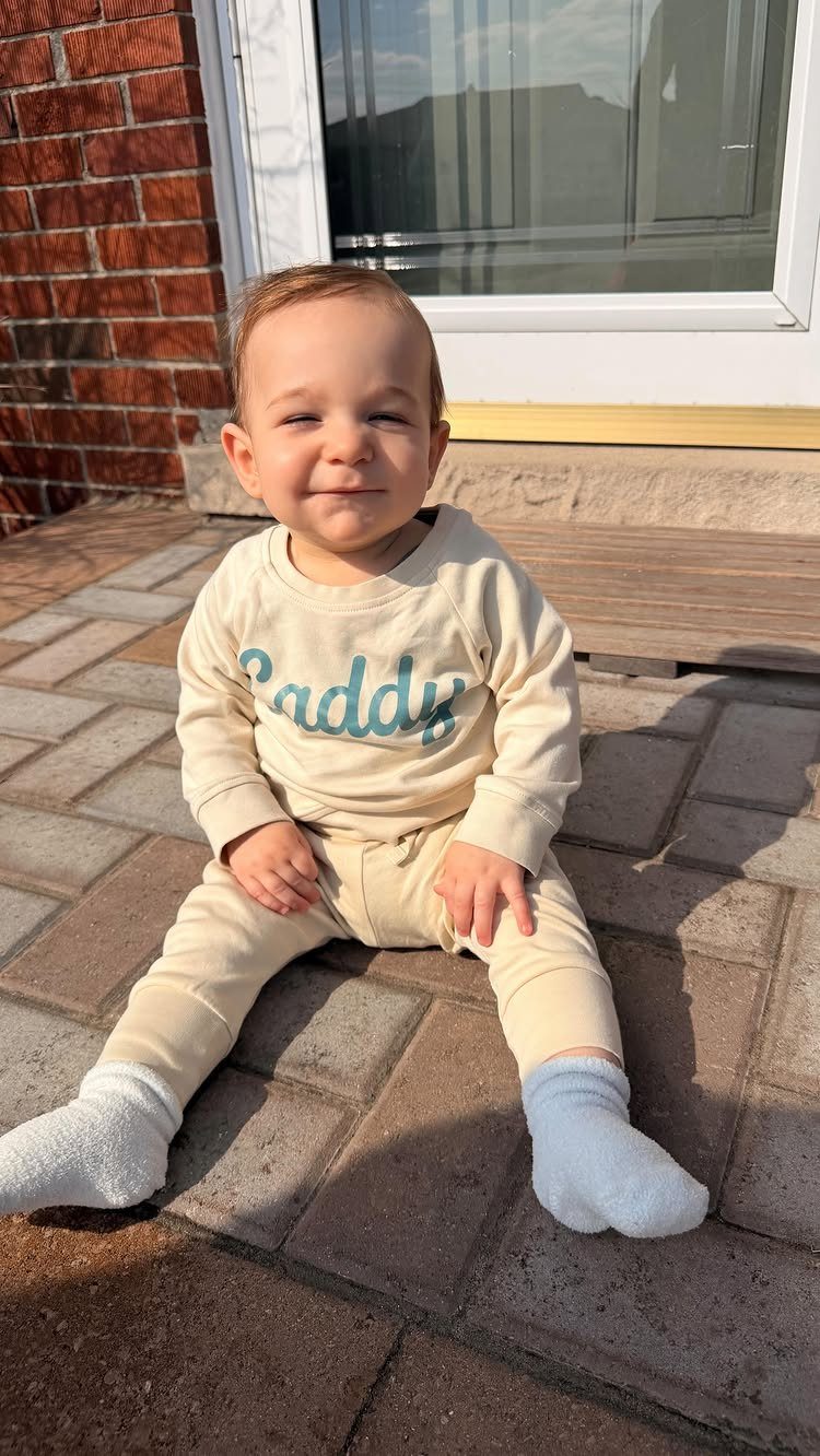Baby sitting on a brick patio wearing cream cotton clothes and white socks.