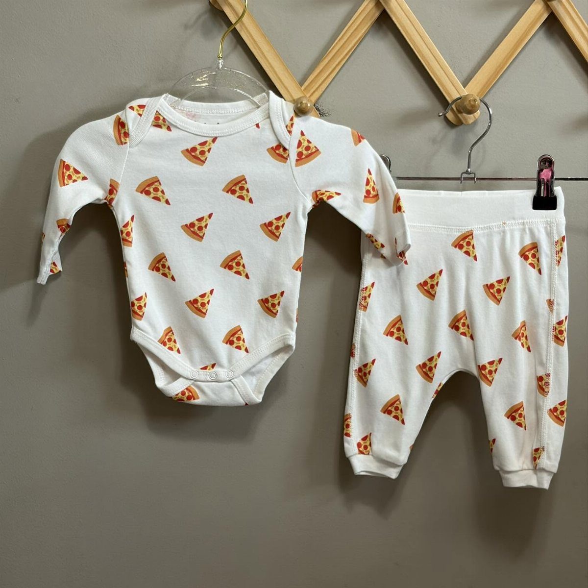 White infant bodysuit and pants with pizza slice prints hanging on hangers.