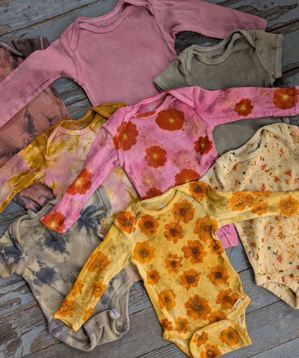 Infant bodysuits in muted natural tones with floral and tie-dye patterns on wood.