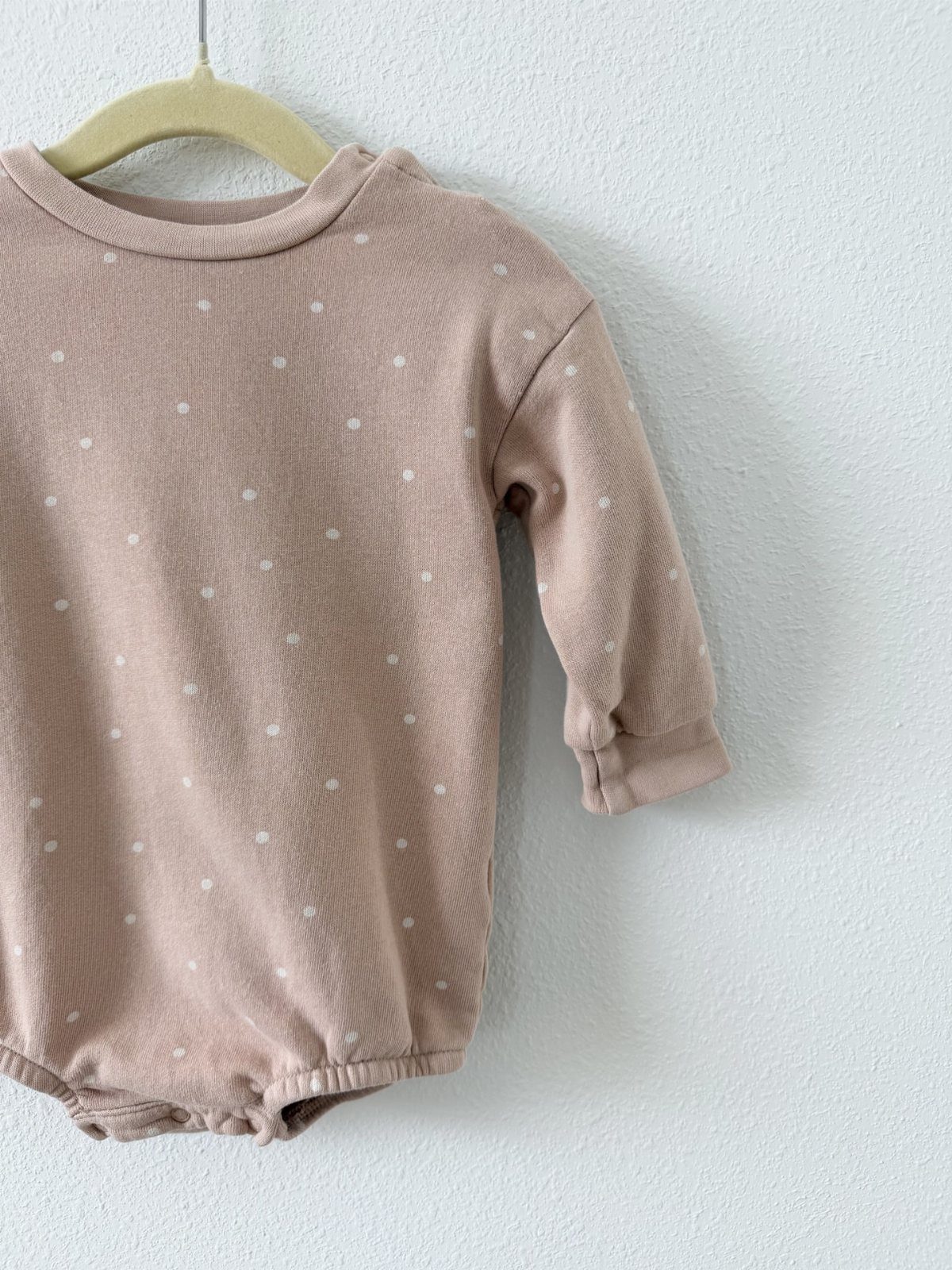 Dusty rose infant bodysuit with white polka dots hanging on a wooden hanger.