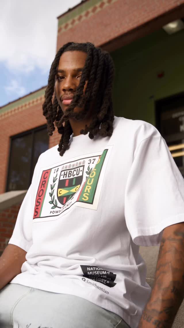 African American man wearing a white HBCU graphic t-shirt and light-colored pants outdoors.