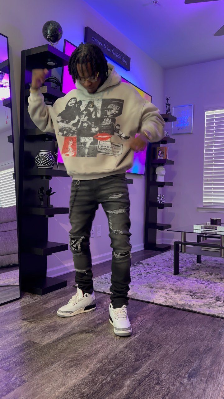 Young Black man wearing a beige graphic hoodie, ripped black jeans, and white sneakers.