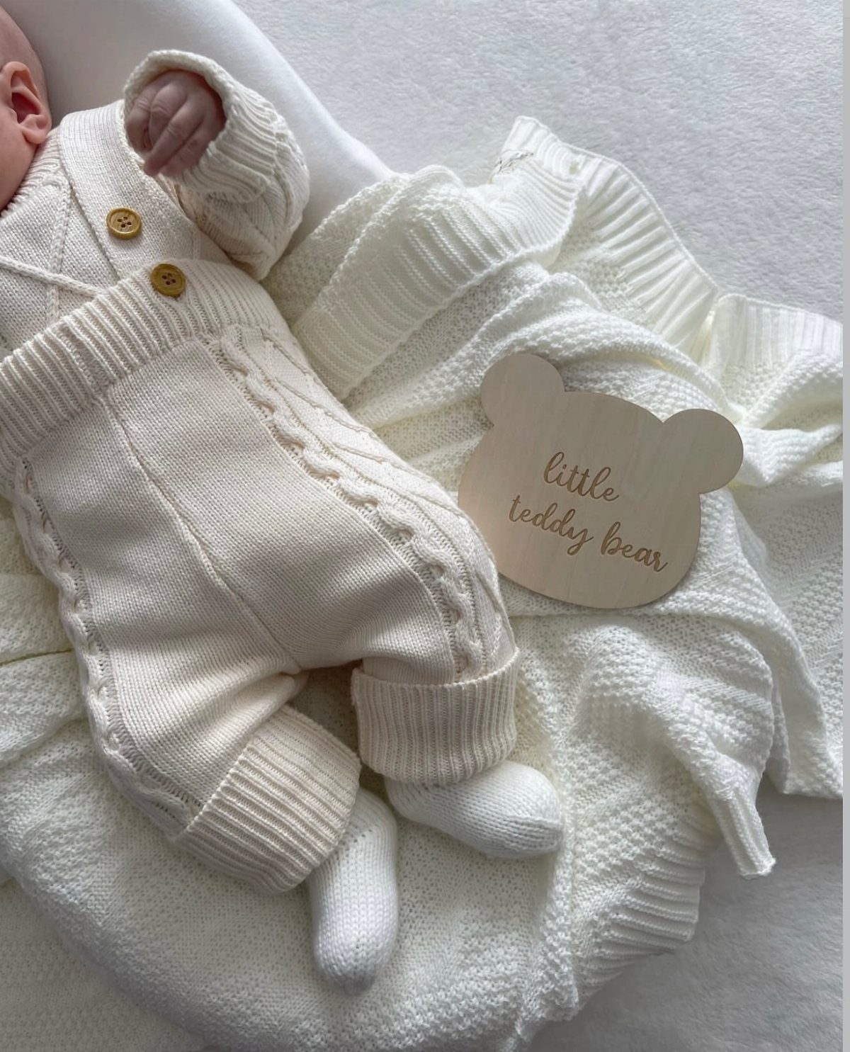 Newborn baby in cream cable-knit outfit lying on white blanket with bear plaque.