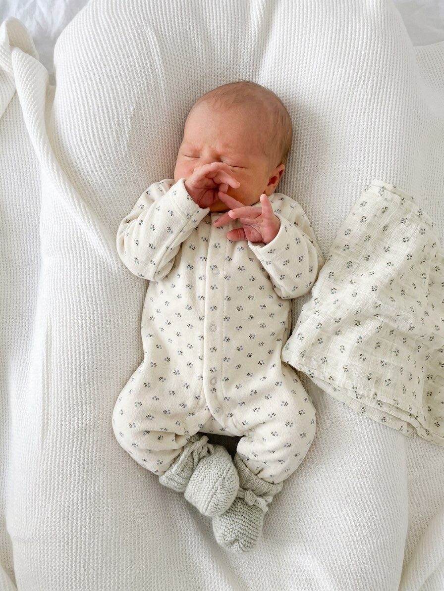 Newborn baby in a white floral print sleeper and grey booties on white bedding
