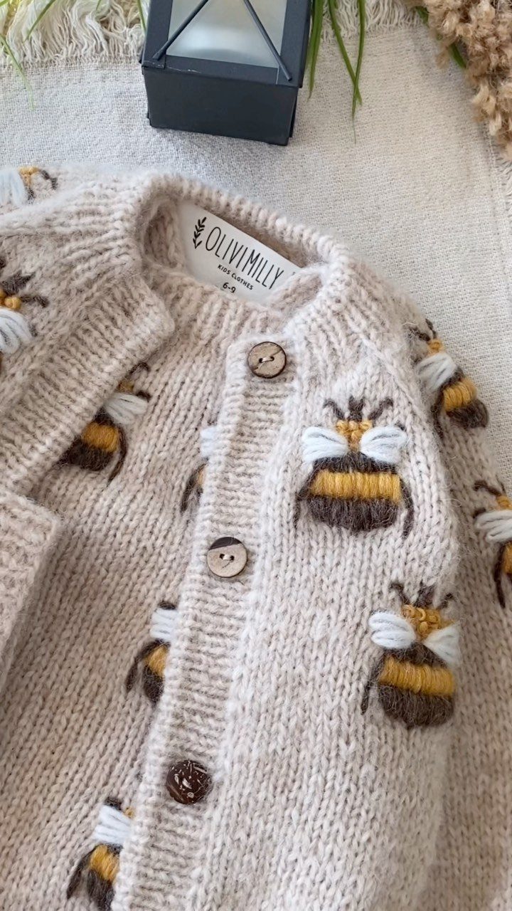Beige knitted baby cardigan featuring embroidered bees and brown buttons on white fabric.