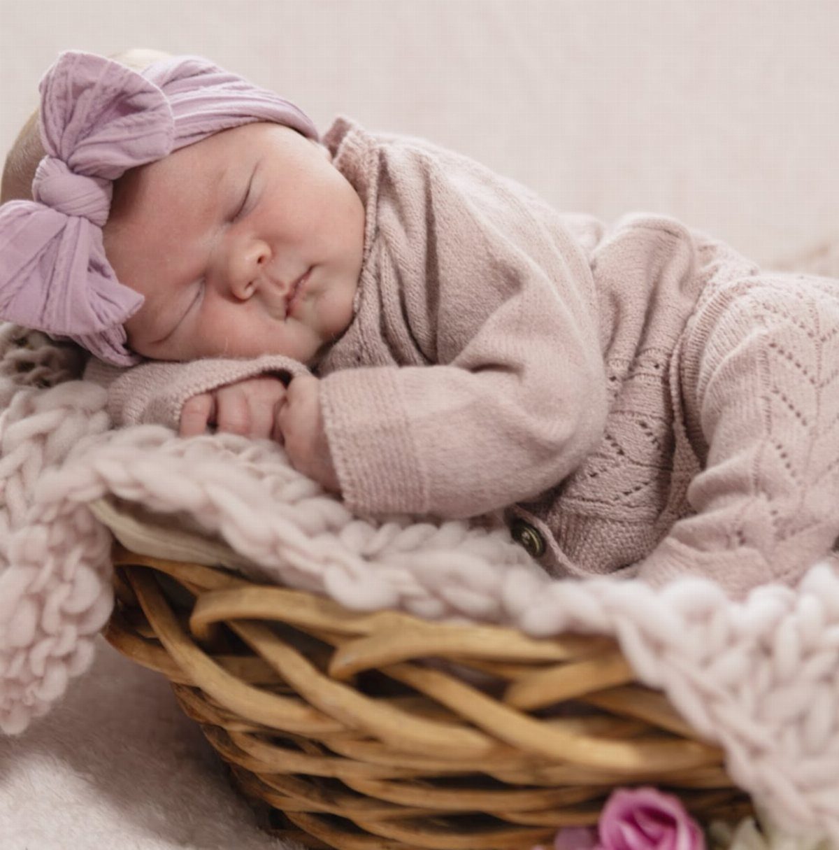 Sleeping newborn baby in a mauve knit outfit and bow headband in a basket