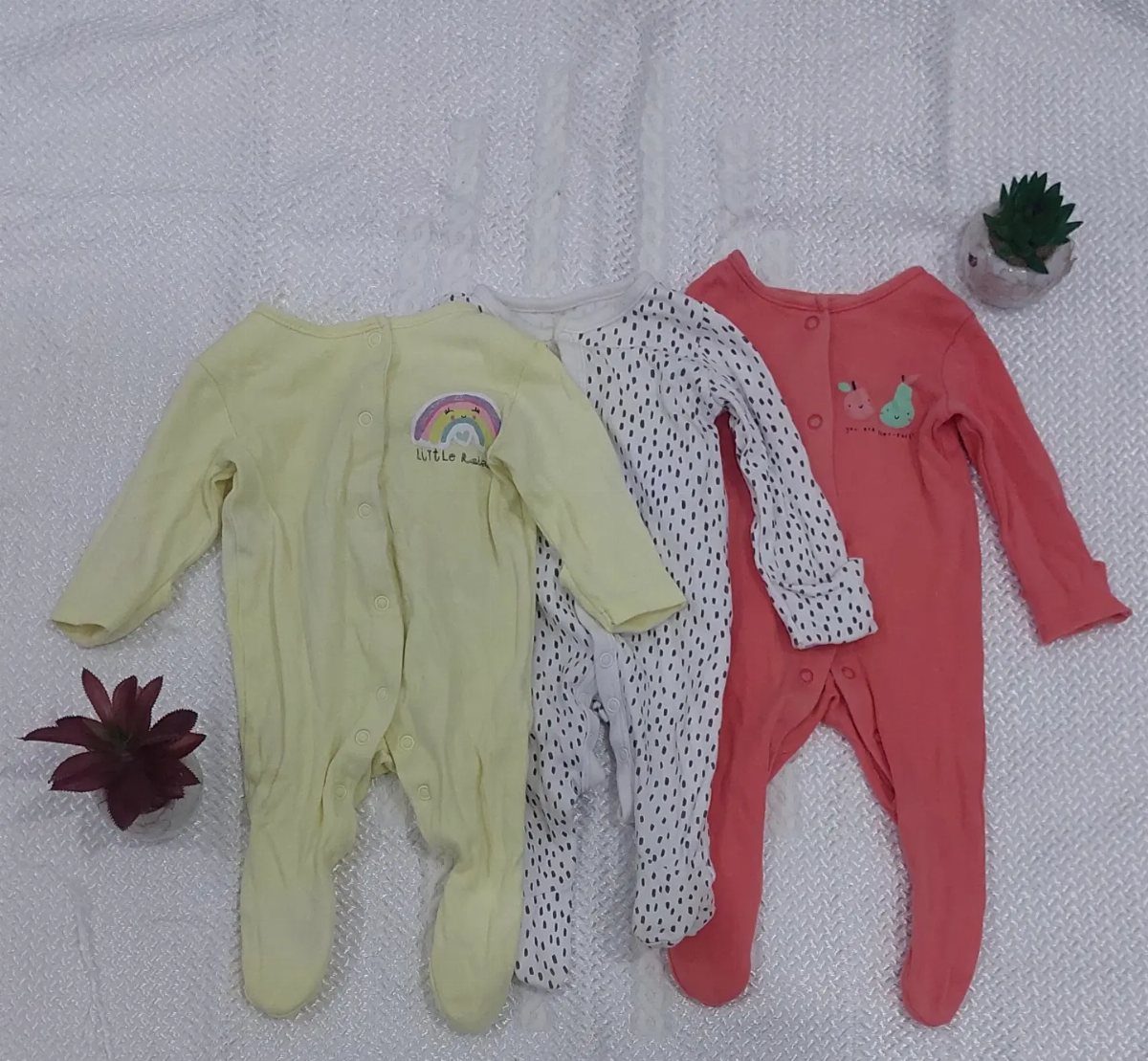 Three newborn footed pajamas in yellow, speckled white, and coral on white fabric.