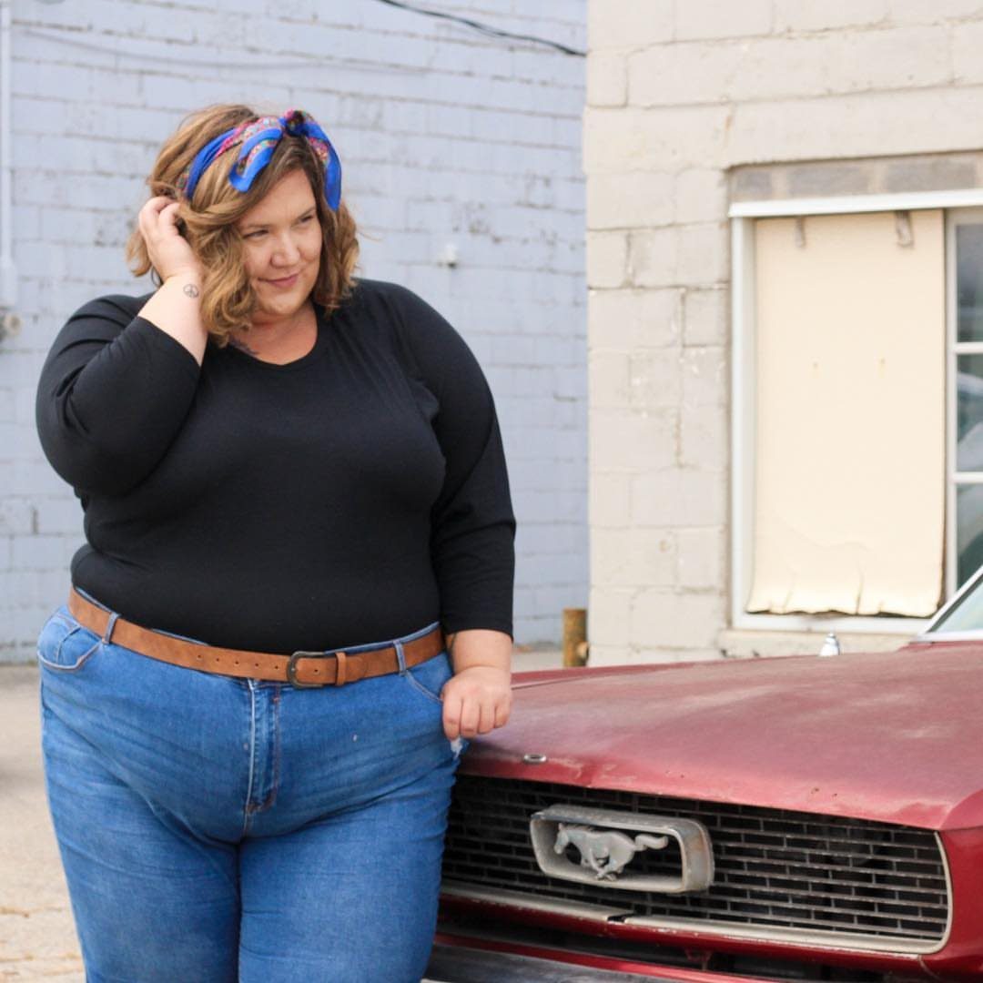 Plus-size woman in black top and blue jeans leaning on red vintage car.
