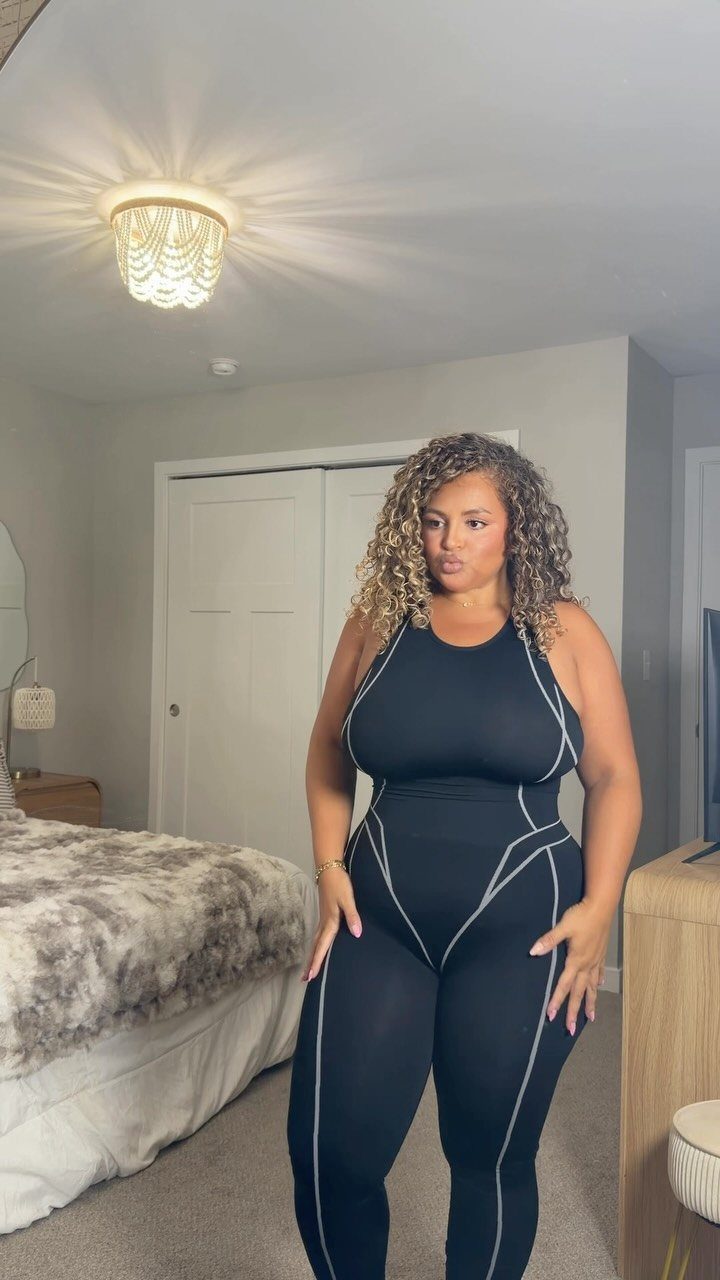Plus-size woman wearing a black sleeveless jumpsuit with white piping in a bedroom.