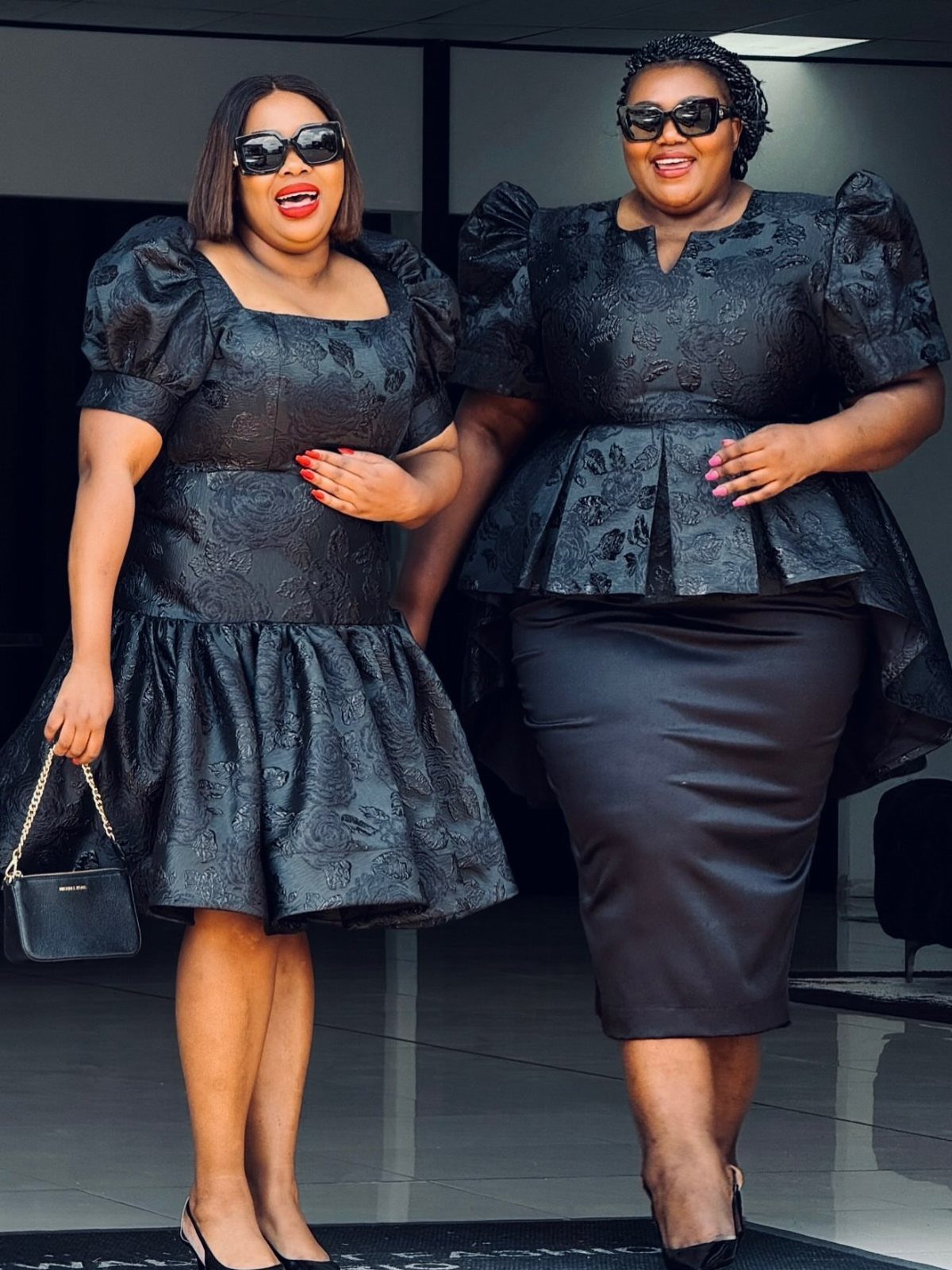 Two plus-size women posing in elegant, structured black dresses and sunglasses indoors.