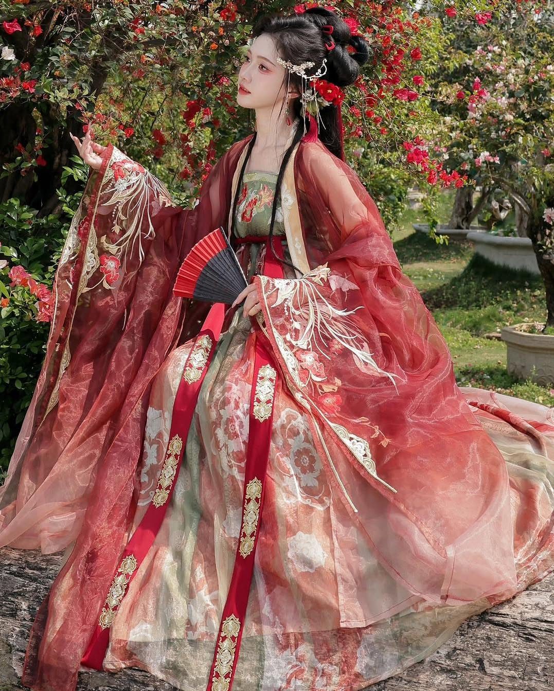 Woman in red and gold Hanfu holding a fan in a garden setting