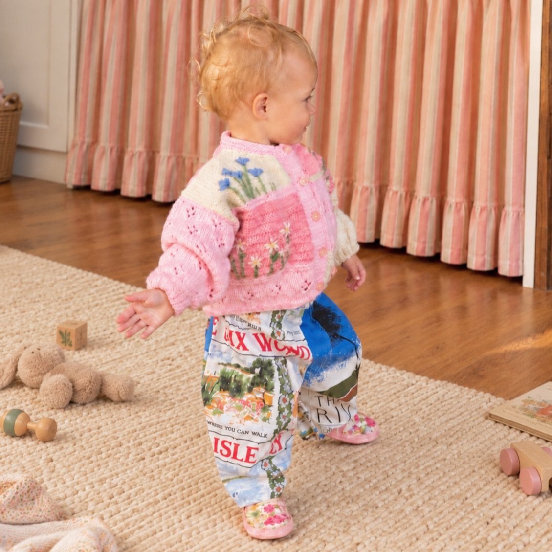 Toddler wearing a pink floral knitted cardigan and printed pants on a rug
