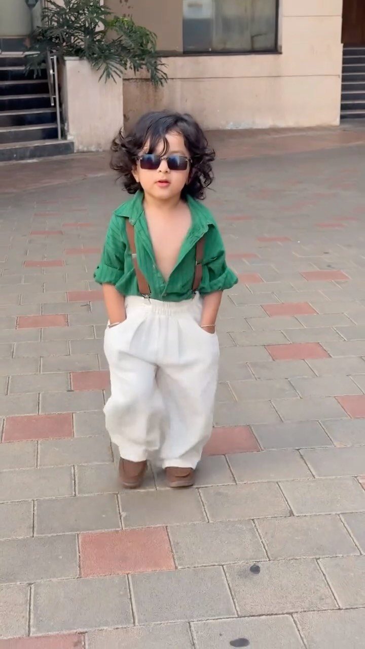 Toddler wearing a green shirt, white trousers, and sunglasses on a tiled walkway