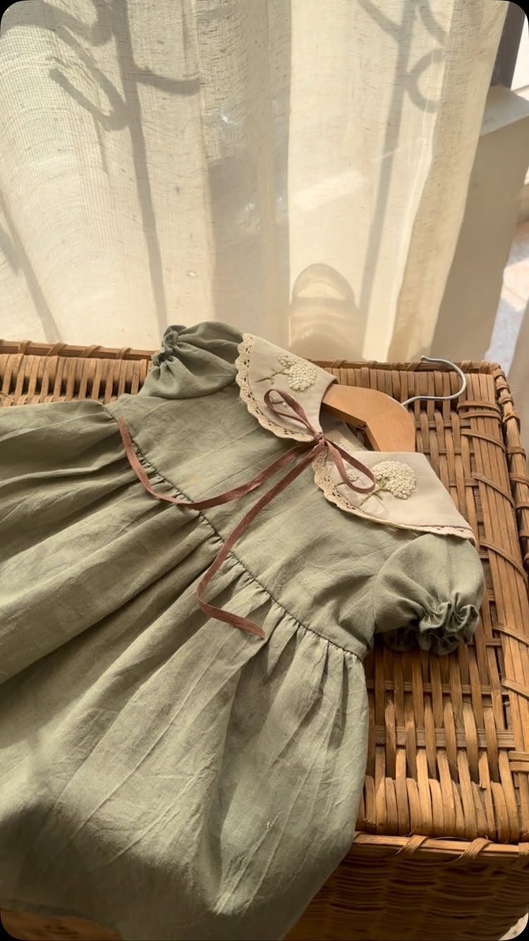Sage green linen baby dress with lace collar on a wicker background