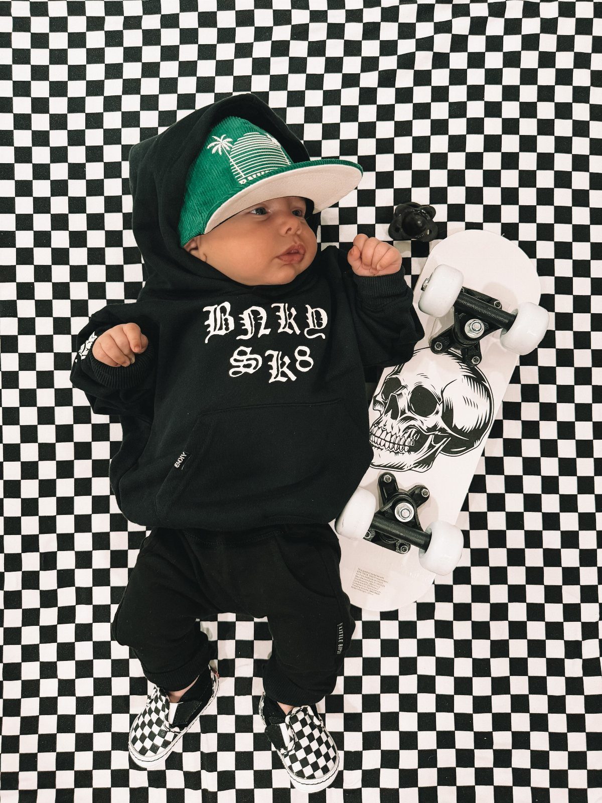 Edgy Infant Clothing Ideas: 6 Looks from Skater to Goth 4 Infant in black streetwear and checkered shoes lying next to a skull skateboard.