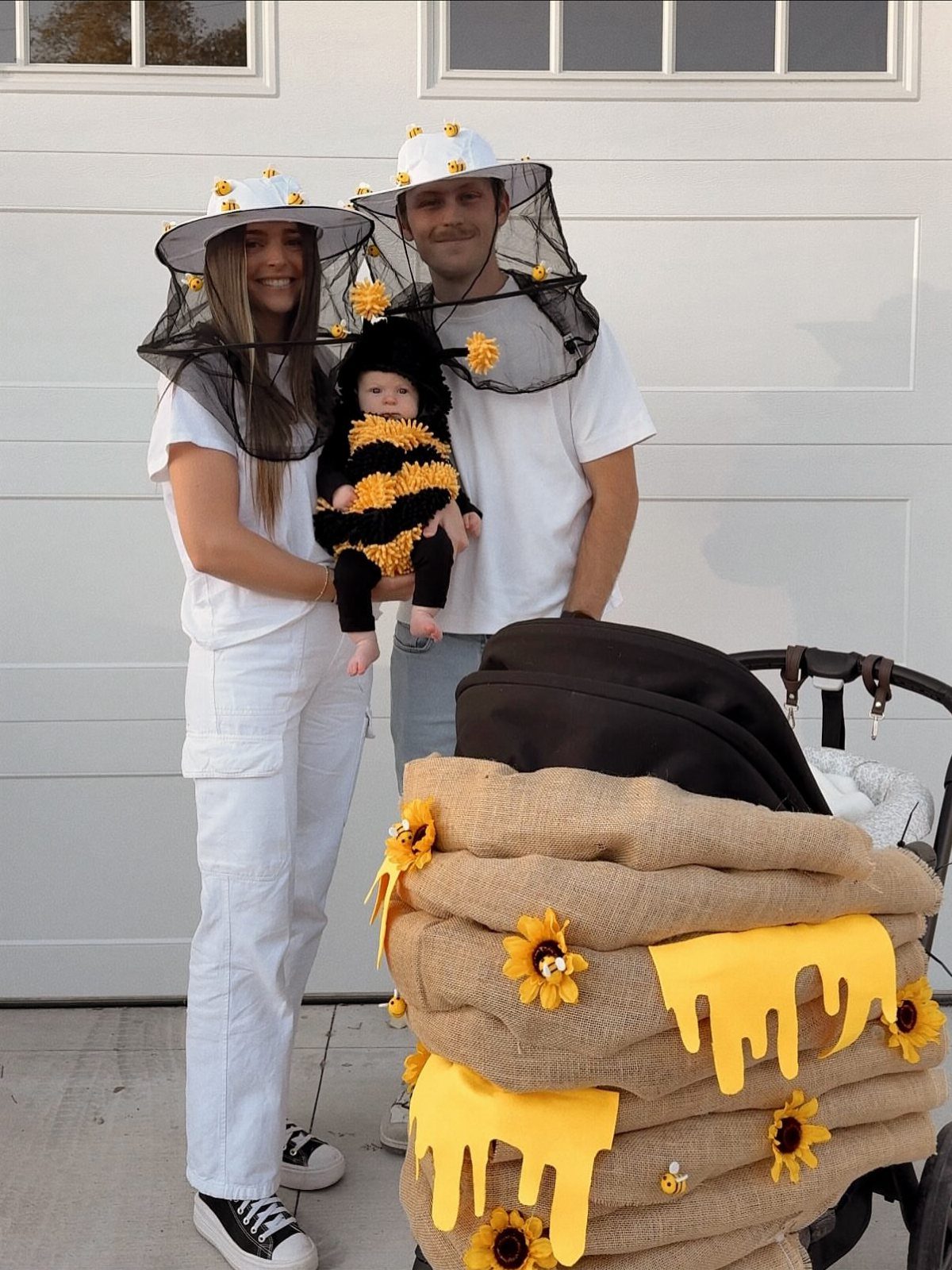 Parents dressed as beekeepers holding a baby in a bee costume next to a honey-pot stroller