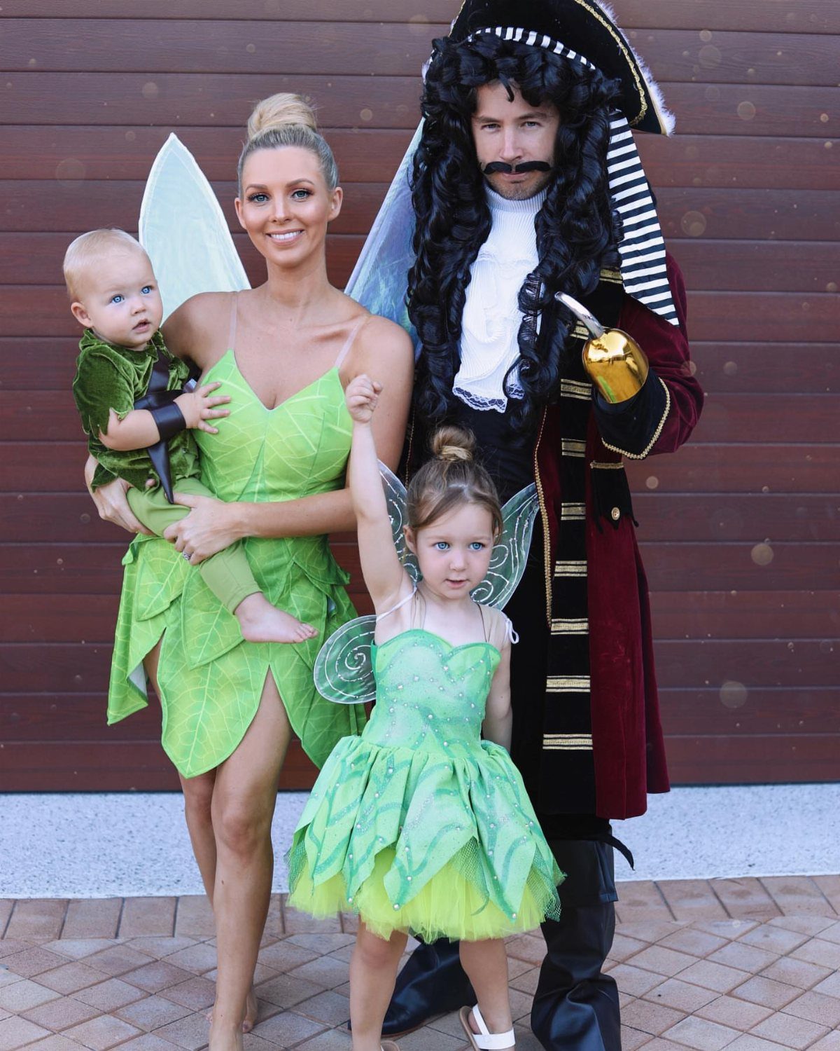 Family in fairy and pirate costumes posing together outdoors against a brown wall.