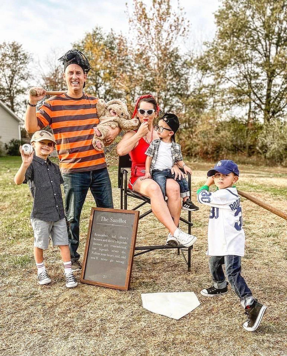 Family in baseball costumes posing outdoors with a newborn and a chalkboard sign.