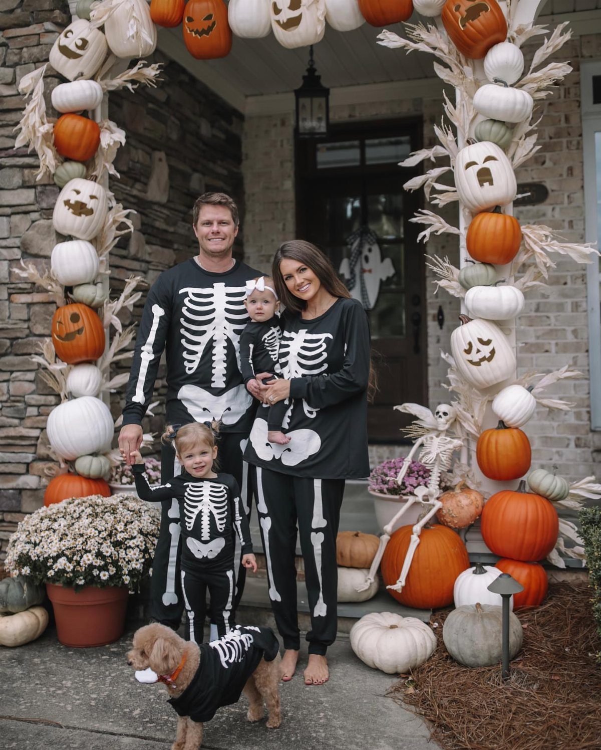 Family and dog wearing matching black skeleton pajamas posing by pumpkin decorations.
