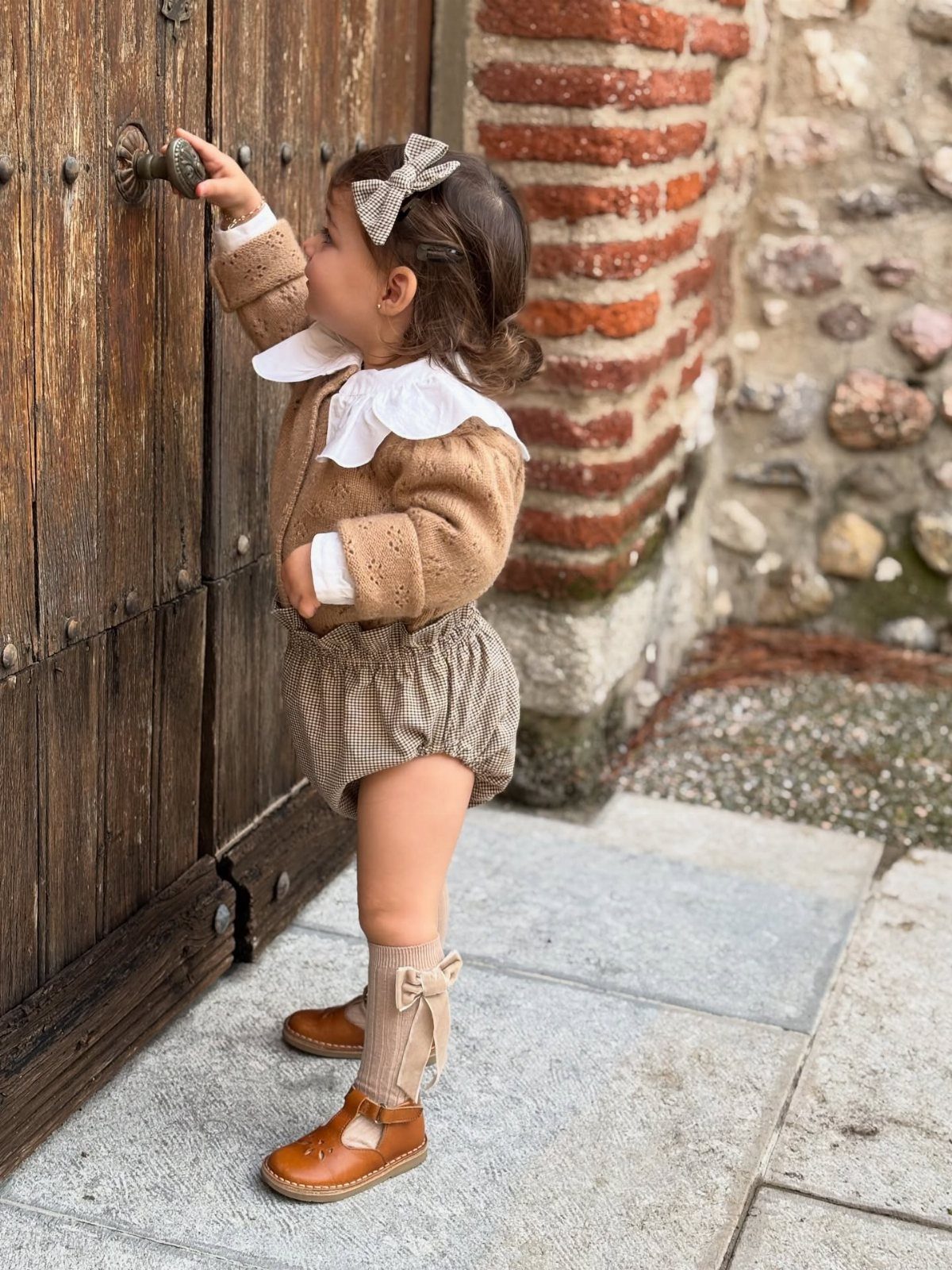 Young child in knit cardigan and checkered bloomers standing by a wooden door.