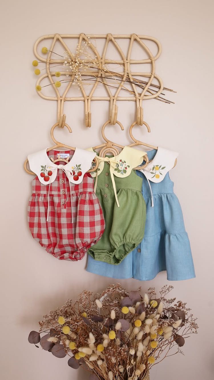 Three infant outfits with embroidered collars hanging on a rattan wall rack.