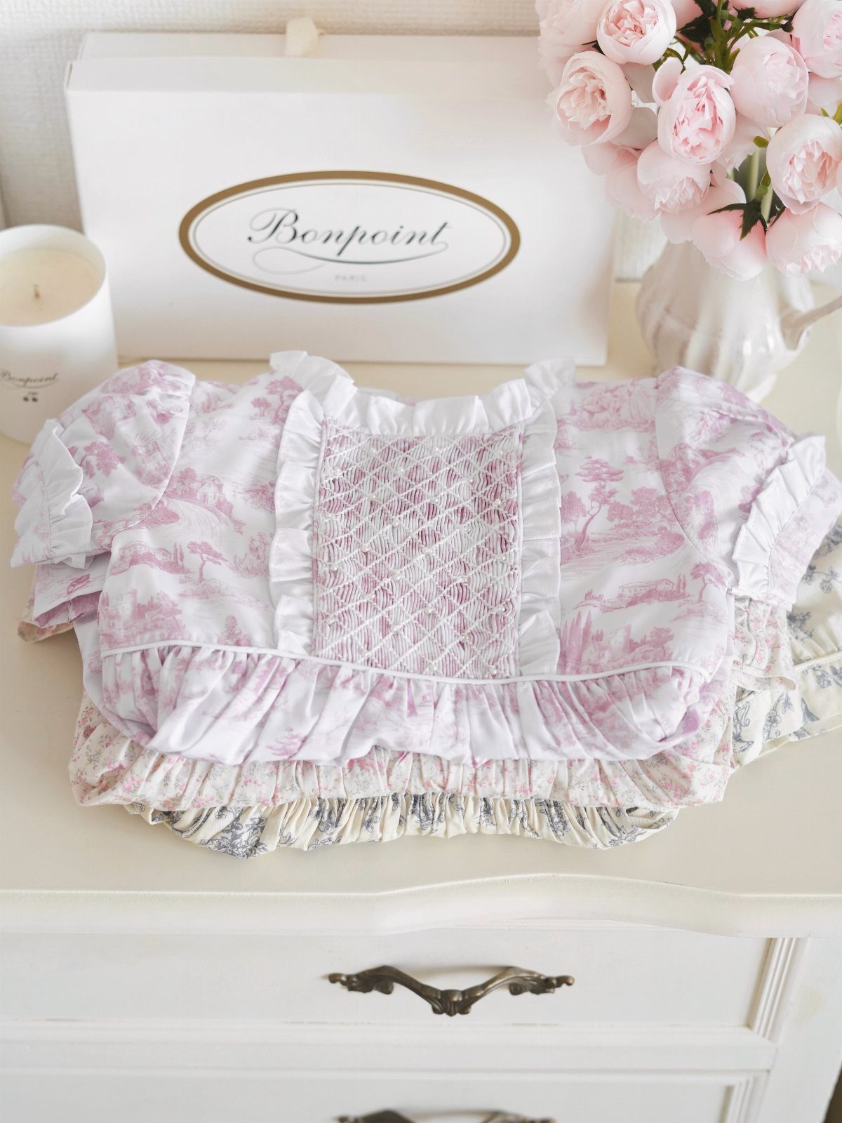 Smocked baby dresses with pink floral patterns stacked on a white dresser.