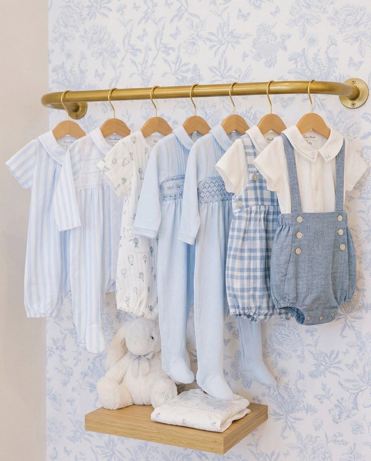 Light blue and white infant rompers hanging on a gold rod against floral wallpaper.