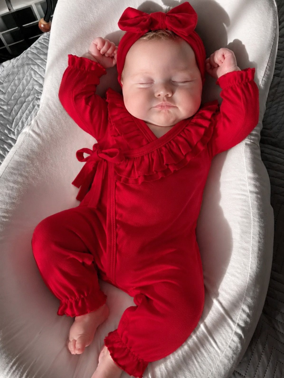 Sleeping infant in a red ruffled romper and matching bow headband on white lounger