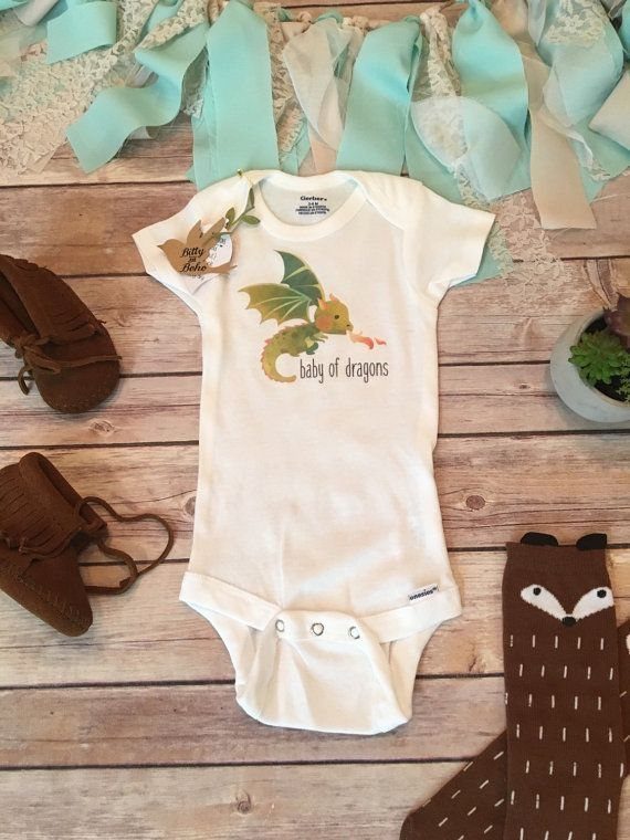 White newborn onesie with green dragon and baby of dragons text on wood.