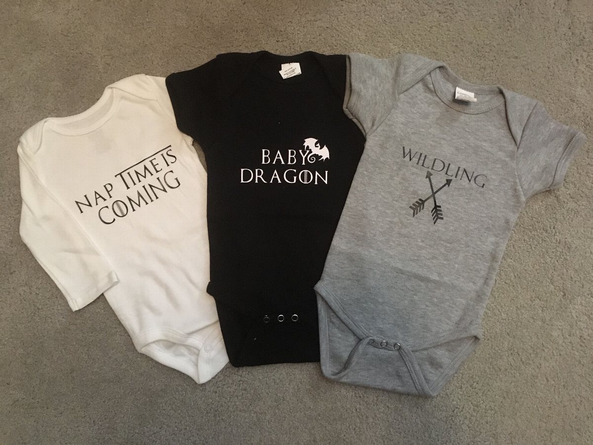 Three infant bodysuits with Game of Thrones themed text and graphics on a grey floor.
