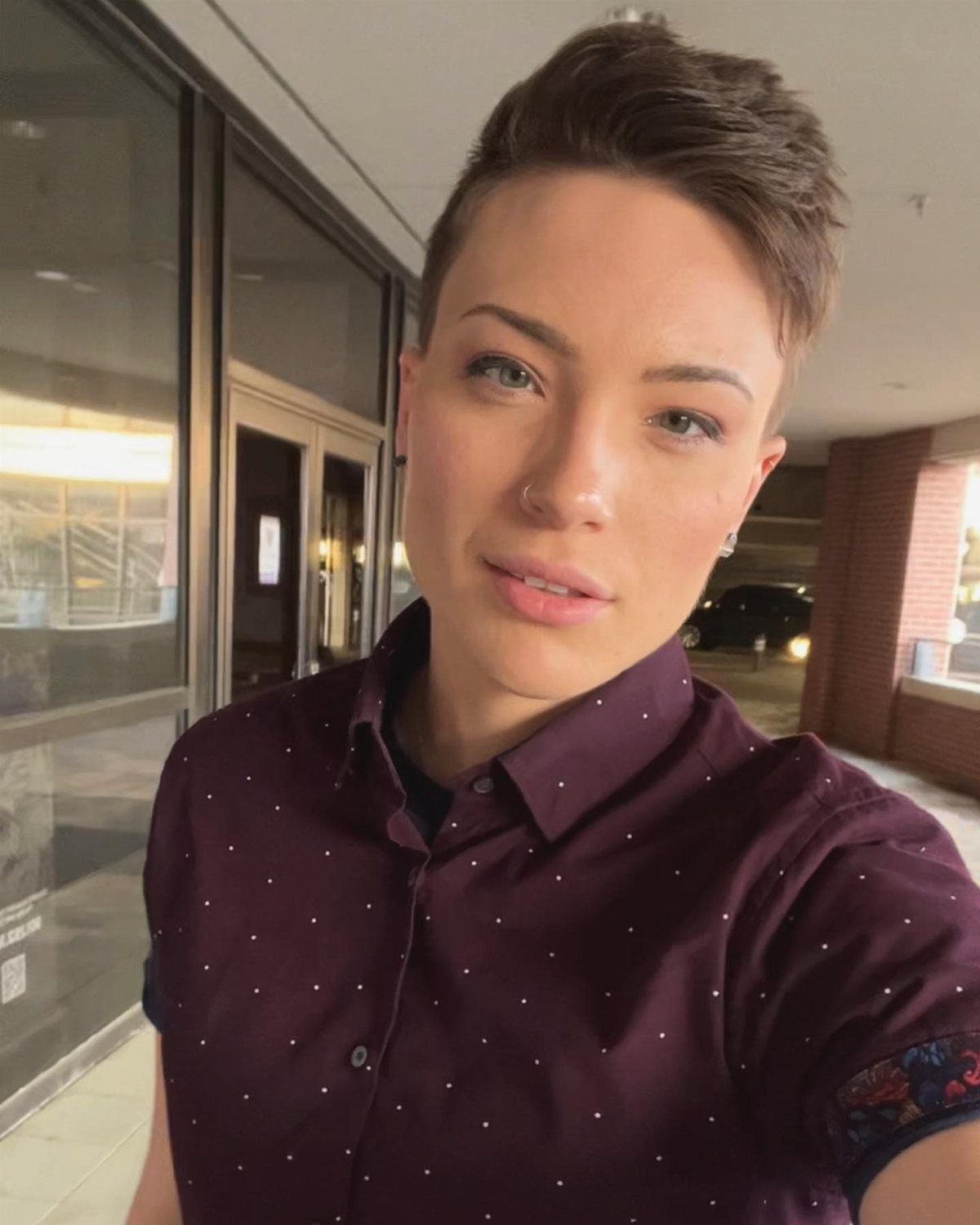 15 Gender Neutral Clothing Ideas for Everyday Style 1 Person with short hair wearing a maroon dotted button-up shirt in a hallway.