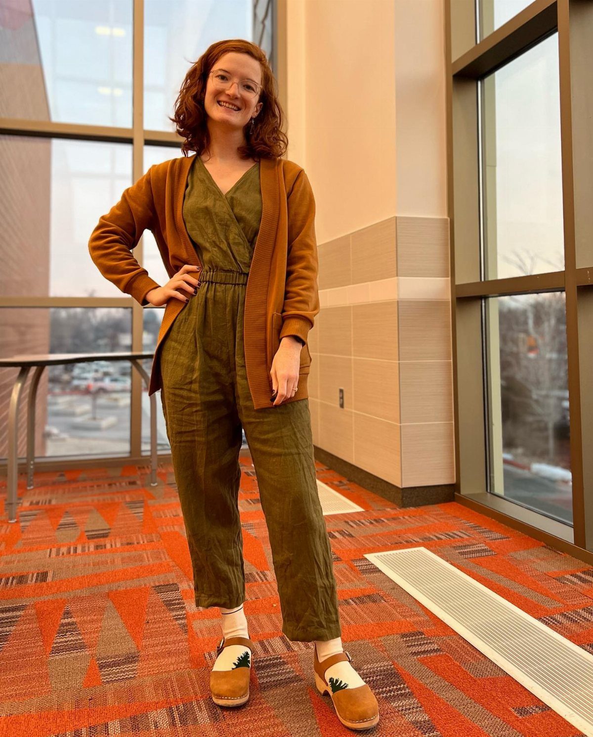 15 Gender Neutral Clothing Ideas for Everyday Style 12 Person wearing an olive green jumpsuit and mustard cardigan standing in a bright room.