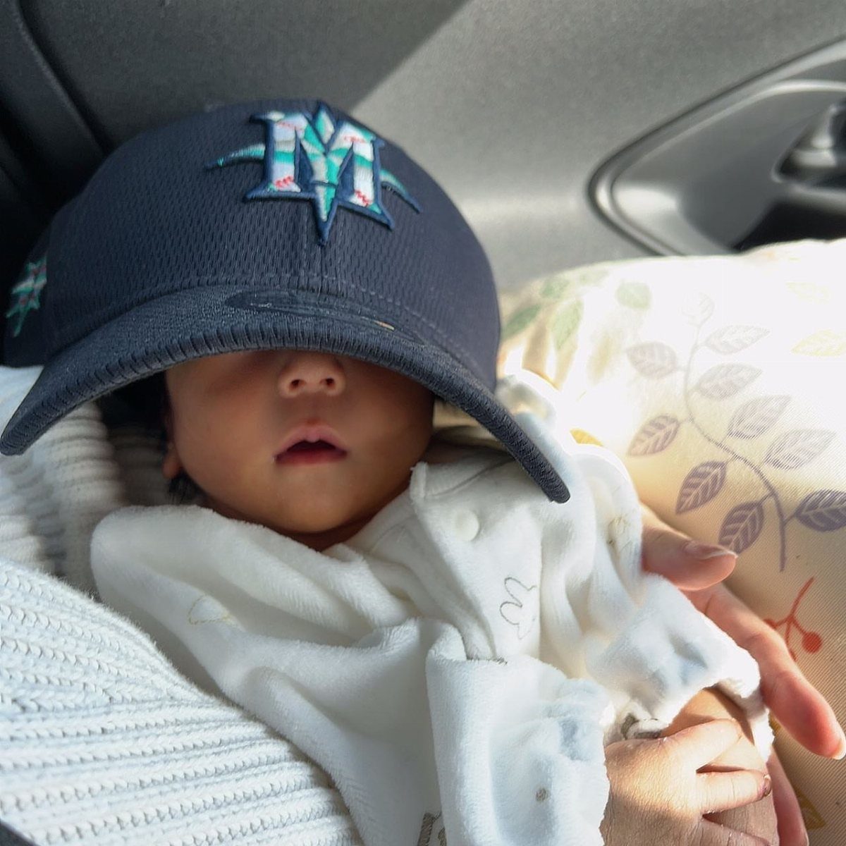 Baby wearing a navy blue Mariners baseball cap and white clothing in a car.