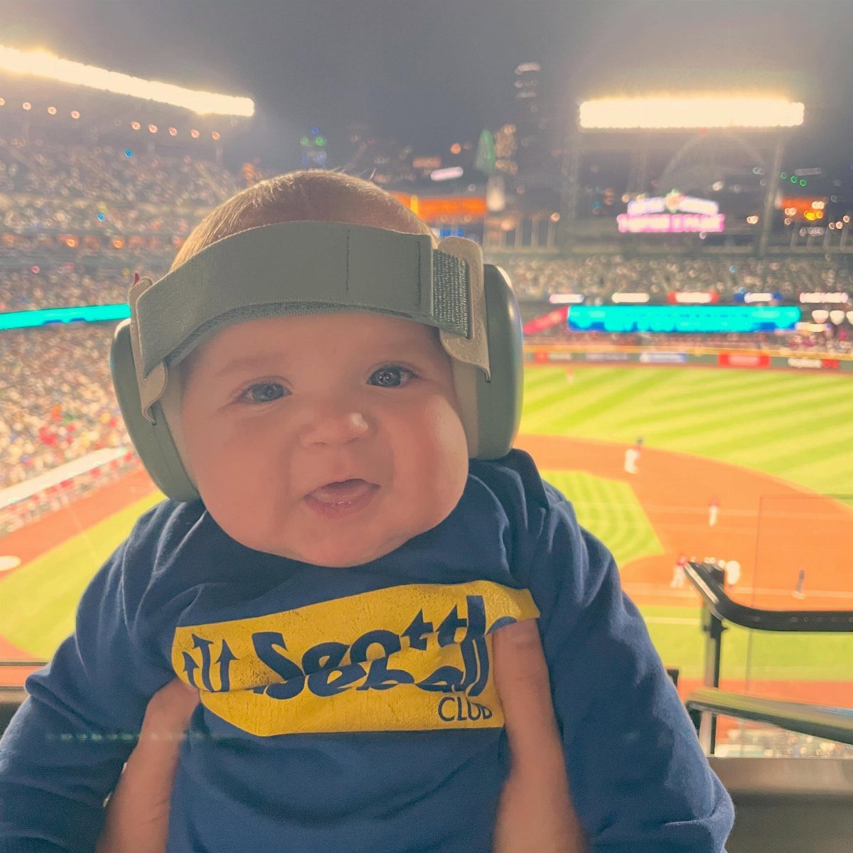Baby wearing a blue Seattle Mariners sweatshirt and green headphones at a stadium.
