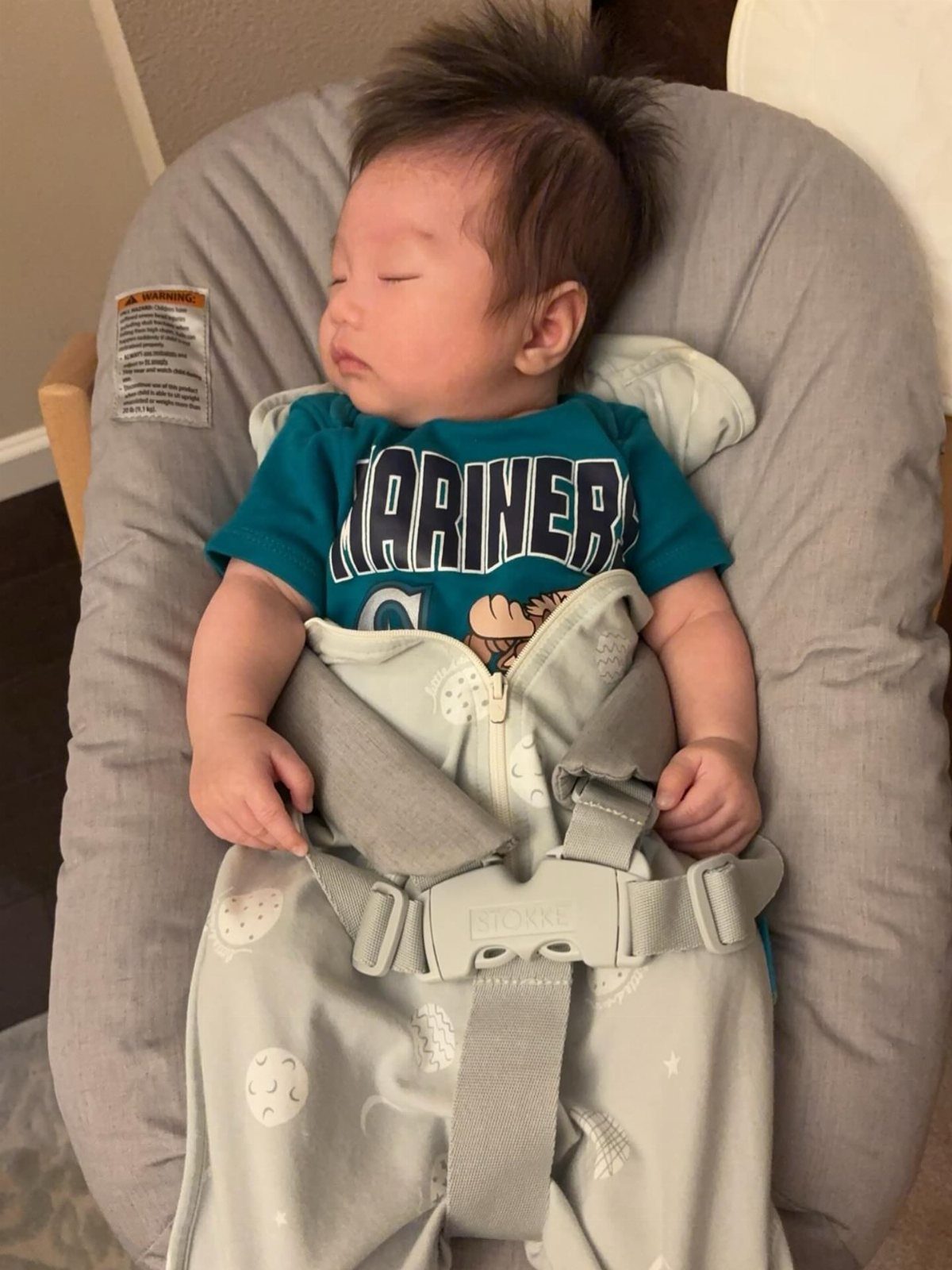 Sleeping infant wearing a teal Mariners short-sleeve bodysuit in a grey baby seat.
