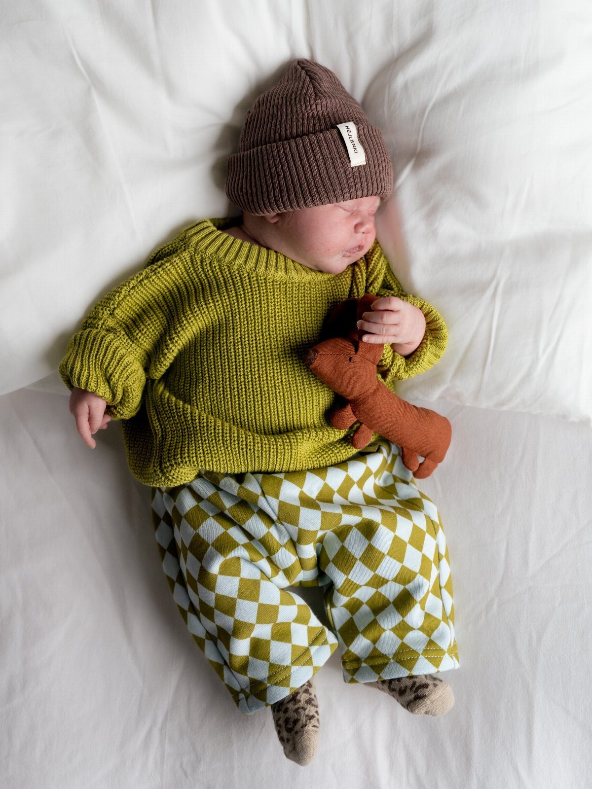 Sleeping baby wearing a green knit sweater and checkered pants on white bedding.