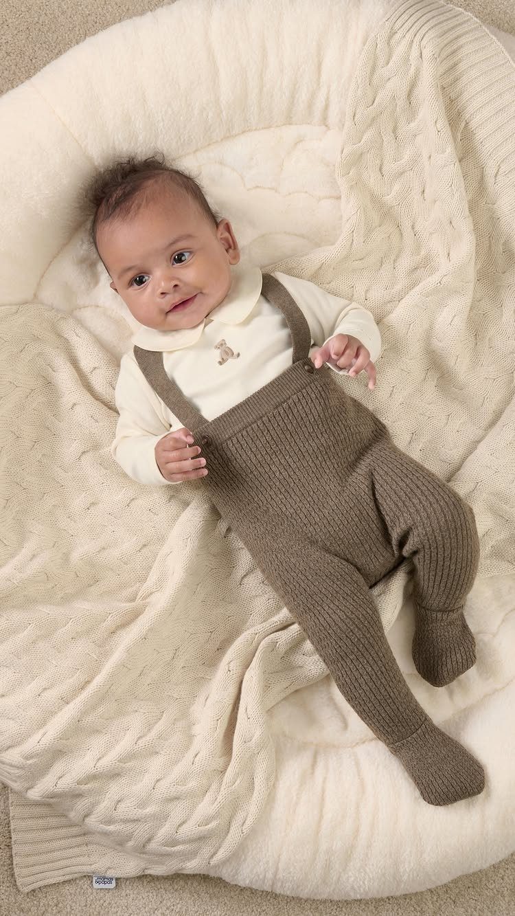Baby wearing brown knitted overalls and white shirt on a cream cable-knit blanket