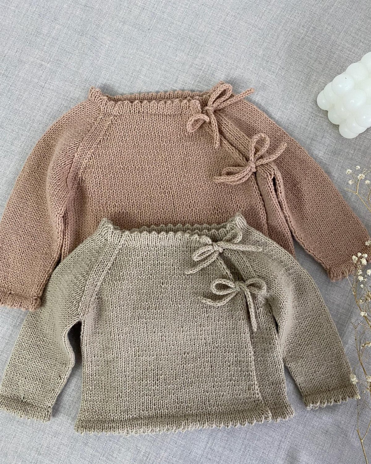 Two hand-knit baby sweaters in dusty rose and sage green on grey fabric.