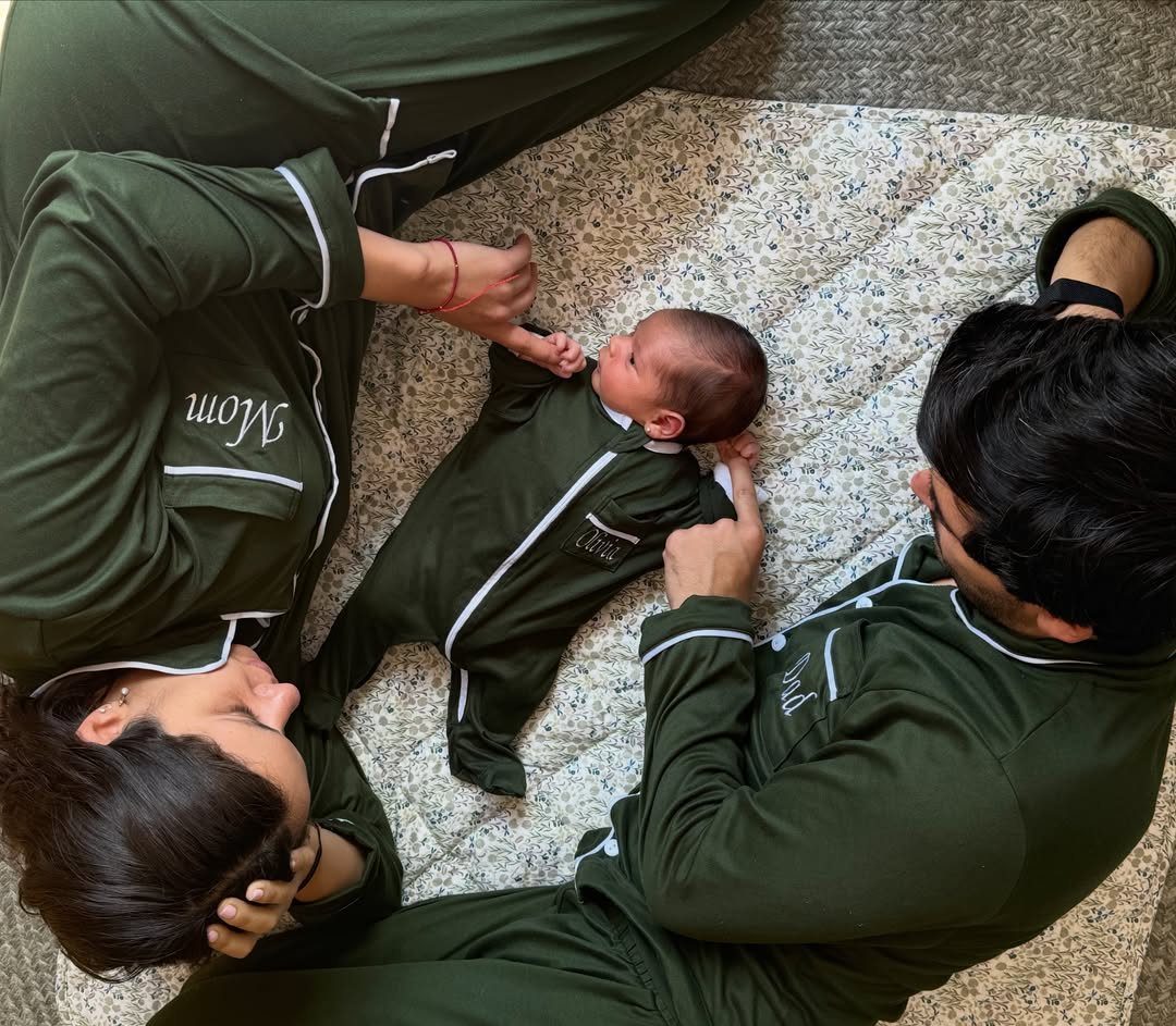 Parents and newborn baby wearing matching dark green pajamas on a white blanket.