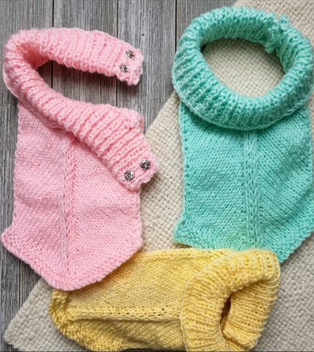 Three knitted baby neck warmers in pink, mint green, and yellow on wood.