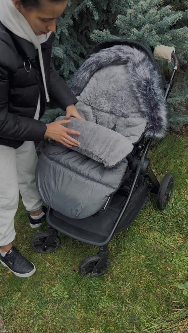 Woman adjusting a grey quilted winter footmuff in a black baby stroller outdoors.
