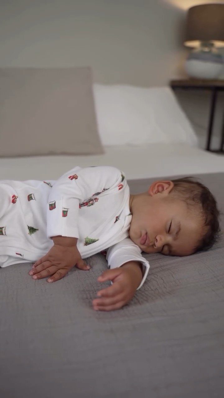 Sleeping baby wearing white pajamas with small Christmas tree prints on a bed.