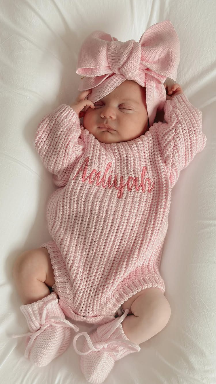 Sleeping newborn baby wearing a pink knitted dress, bow headband, and matching booties.
