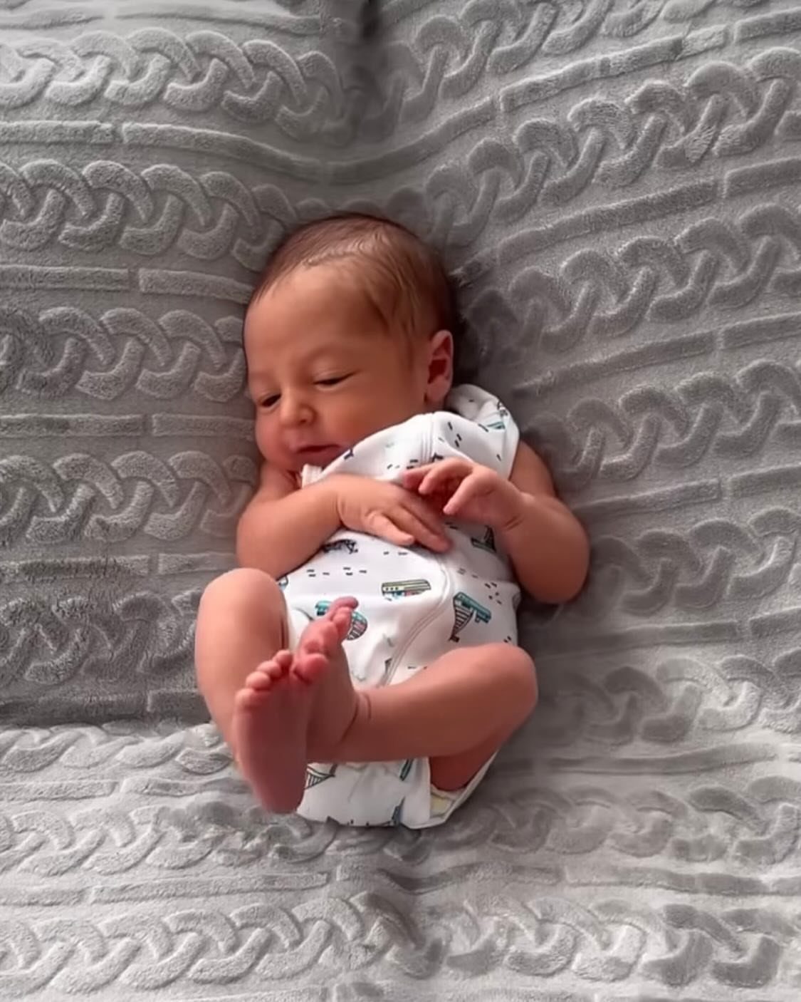 Newborn baby wearing a white patterned bodysuit lying on a grey knit blanket