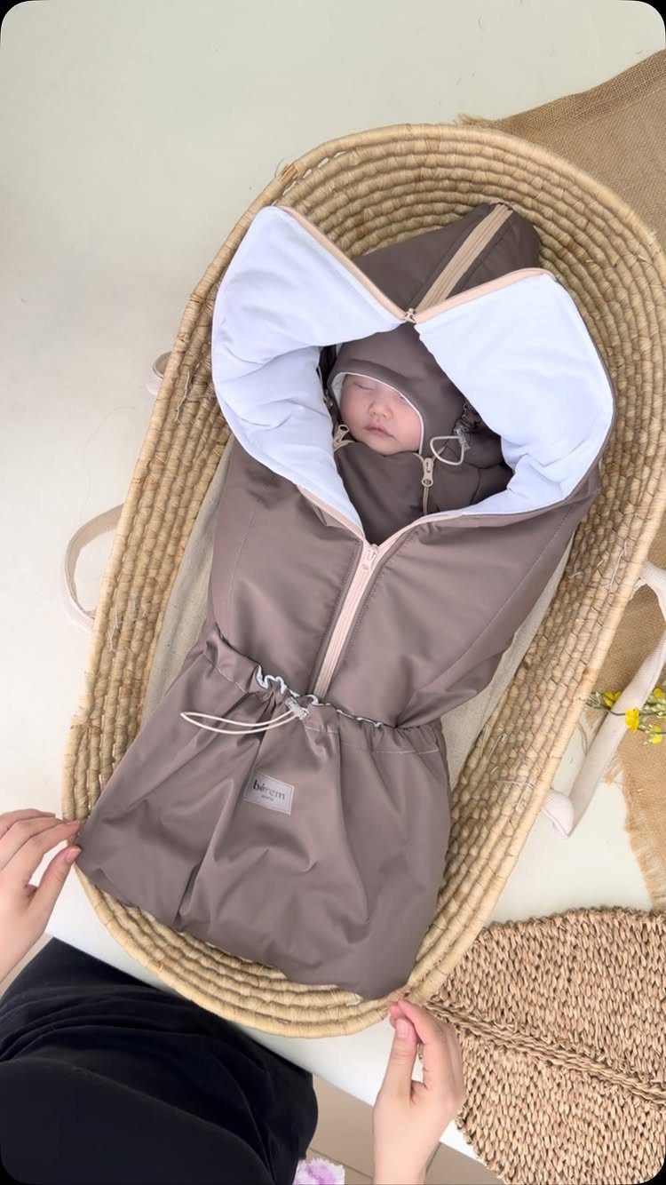 Newborn baby wearing a brown bunting bag lying in a woven wicker bassinet.