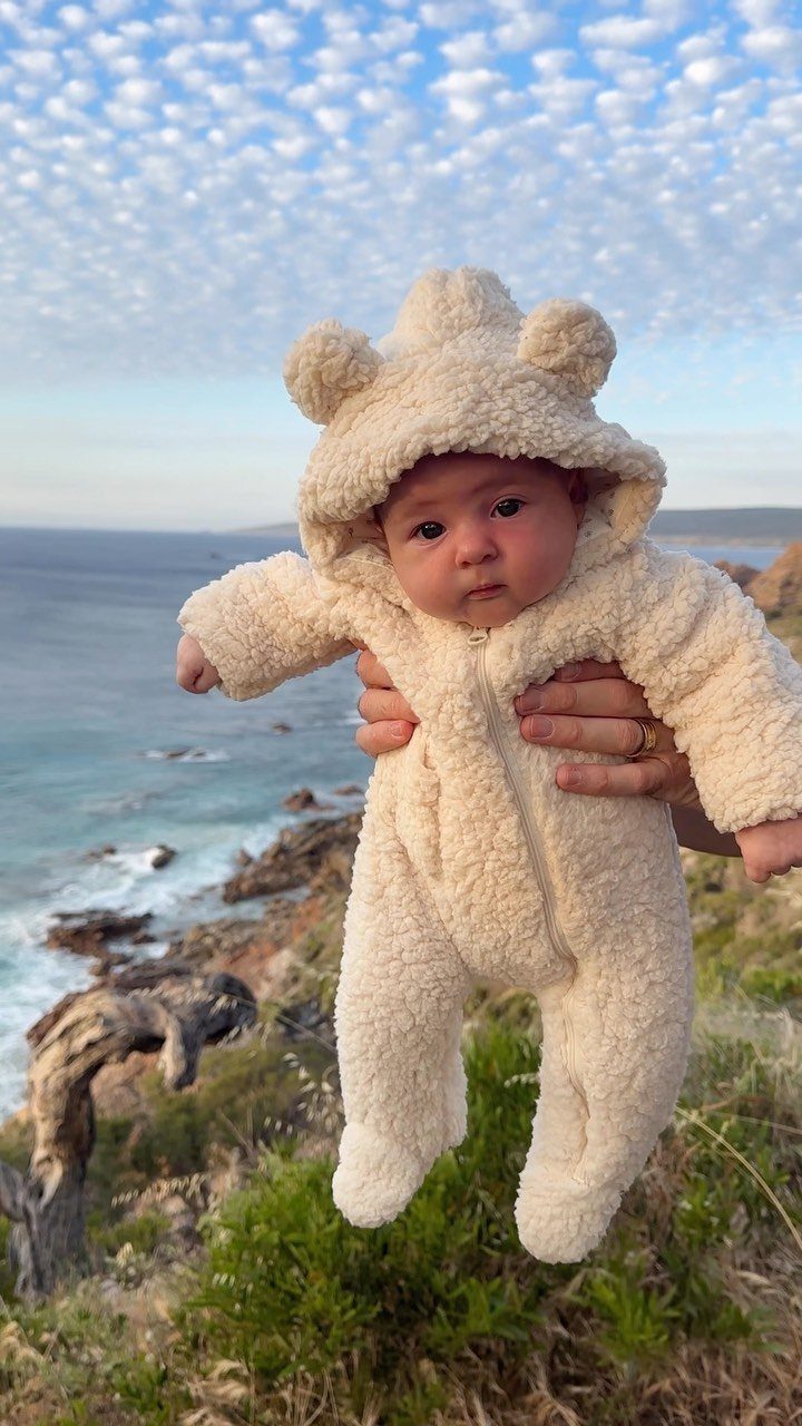 Newborn baby wearing a cream fuzzy bear-eared fleece jumpsuit outdoors by the ocean.
