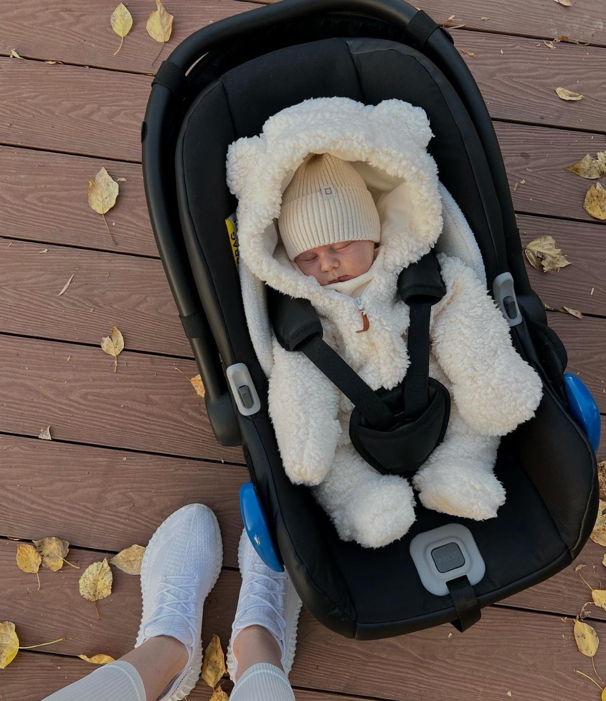 Newborn baby in a white fluffy bear snowsuit and beanie in a car seat.