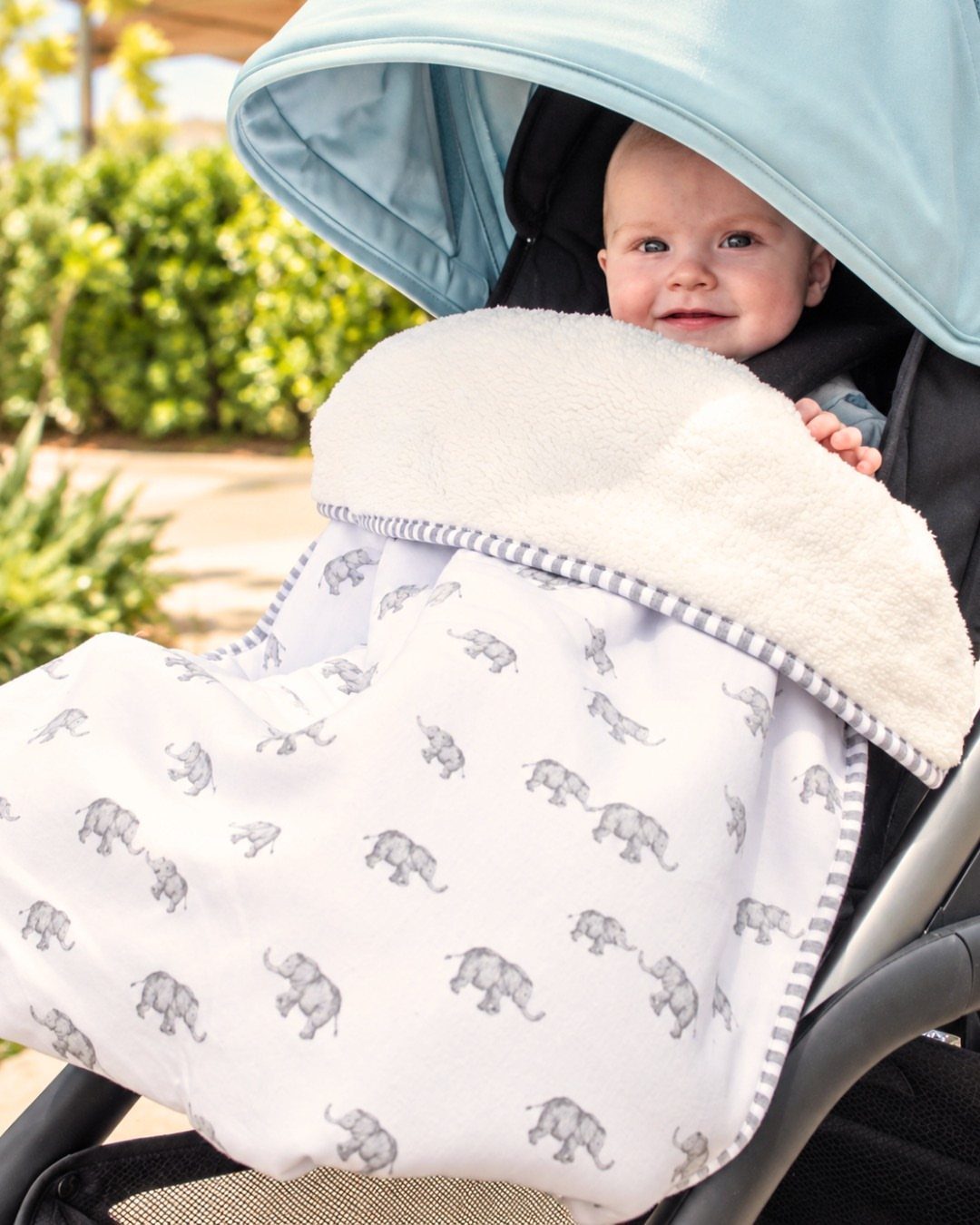Baby in a blue stroller wrapped in a white elephant print warm blanket.